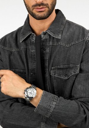 Orologio cronografo d'argento con quadrante bianco, cinturino in acciaio inox e più sub-quadranti. Indossato su un polso sotto una giacca di jeans nera.