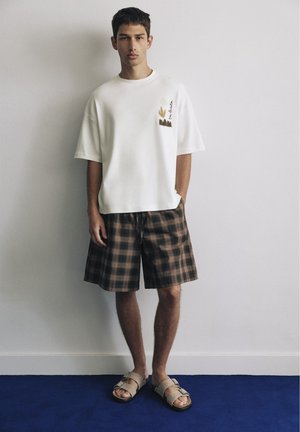 Ung mand står op ad en hvid væg iført en oversized hvid T-shirt, brune ternede shorts og beige sandaler på blå gulv.