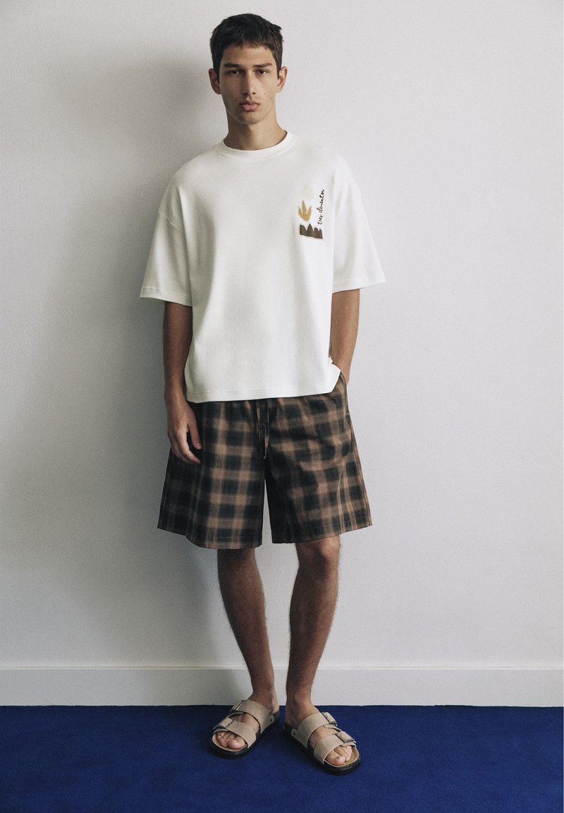 Jeune homme debout contre un mur blanc portant un t-shirt blanc oversize, un short marron à carreaux et des sandales beiges sur un sol bleu.