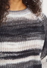 Nahaufnahme einer Person, die einen gestrickten Pullover mit horizontalen Streifen in Grautönen, Weiß und Schwarz trägt und lockige Haare hat.