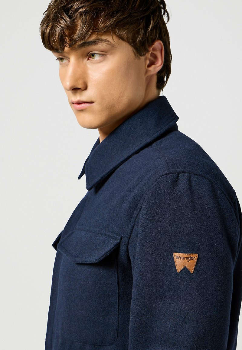 Cappotto di lana navy con alto colletto, dotato di tasca sul petto e di un logo in pelle Wrangler sulla spalla sinistra. Tessuto morbido.
