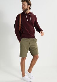 Bordeaux hoodie met witte trekkoorden, olijfgroene shorts en witte sneakers. Inclusief een bruine schoudertas. Casual kleding.