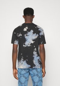 Schwarzes Batik-T-Shirt mit blauen und weißen Mustern, kurzen Ärmeln und Rundhalsausschnitt; getragen mit hellblauen Jeans mit Textdruck.