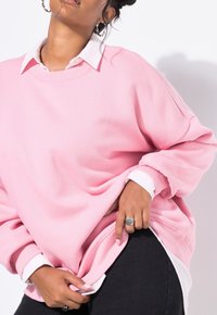 Donna che indossa un felpa oversize rosa sopra una camicia bianca con colletto, pantaloni neri, anello d'argento e capelli mossi con orecchini a cerchio.