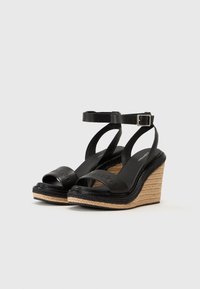Black wedge sandals with an open toe, featuring strappy design and ankle buckle. Woven jute detailing on the wedge heel adds texture.