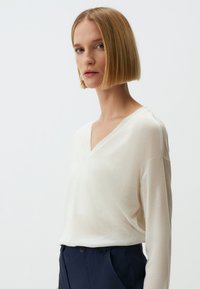 V-hals lange mouwen shirt in een zachte, lichte crèmekleurige stof; relaxte pasvorm met verlaagde schouders; gecombineerd met marineblauwe broeken, minimalistisch ontwerp.