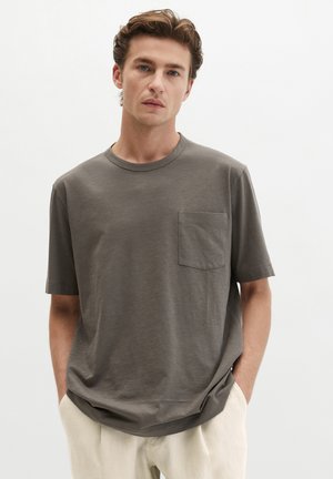 Jonge man draagt een loszittend grijs T-shirt met korte mouwen en borstzakje, gecombineerd met lichtbeige broek, handen in de zakken, neutrale achtergrond.