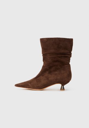 Stivale alla caviglia in suede marrone con un design morbido, punta affilata e tacco sottile. Presenta una texture morbida e pochi dettagli in metallo.