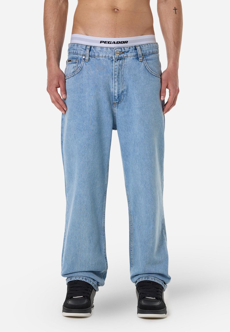 Ανοιχτό μπλε τζιν παντελόνι, σε χαλαρή εφαρμογή, με μέση μέσου ύψους, που διαθέτει παραδοσιακές τσέπες και μεταλλικό κούμπωμα. Φοριέται με μαύρα αθλητικά παπούτσια.