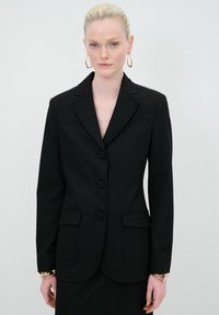 Blazer nero su misura con revers, tre bottoni, due tasche frontali e una texture liscia. Accentato con un bracciale d'oro al polso.