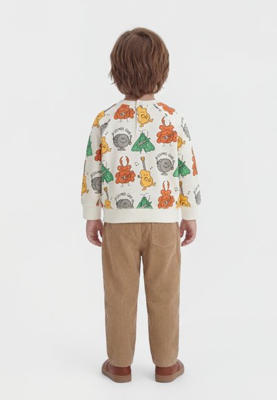 Barn med brunt hår iført en hvid sweatshirt med farverige musikinstrumentfigurer og brune fløjlsbukser, set bagfra.