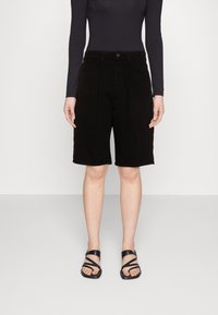 Henkilö, joka pukeutunut mustiin, vyötäröltä korkeavyötäröisiin polvipitkiin shortseihin, pitkähihainen tumma yläosa ja mustat remmityyliset tasapohjasandaalit, seisomassa yksivärisen taustan edessä.