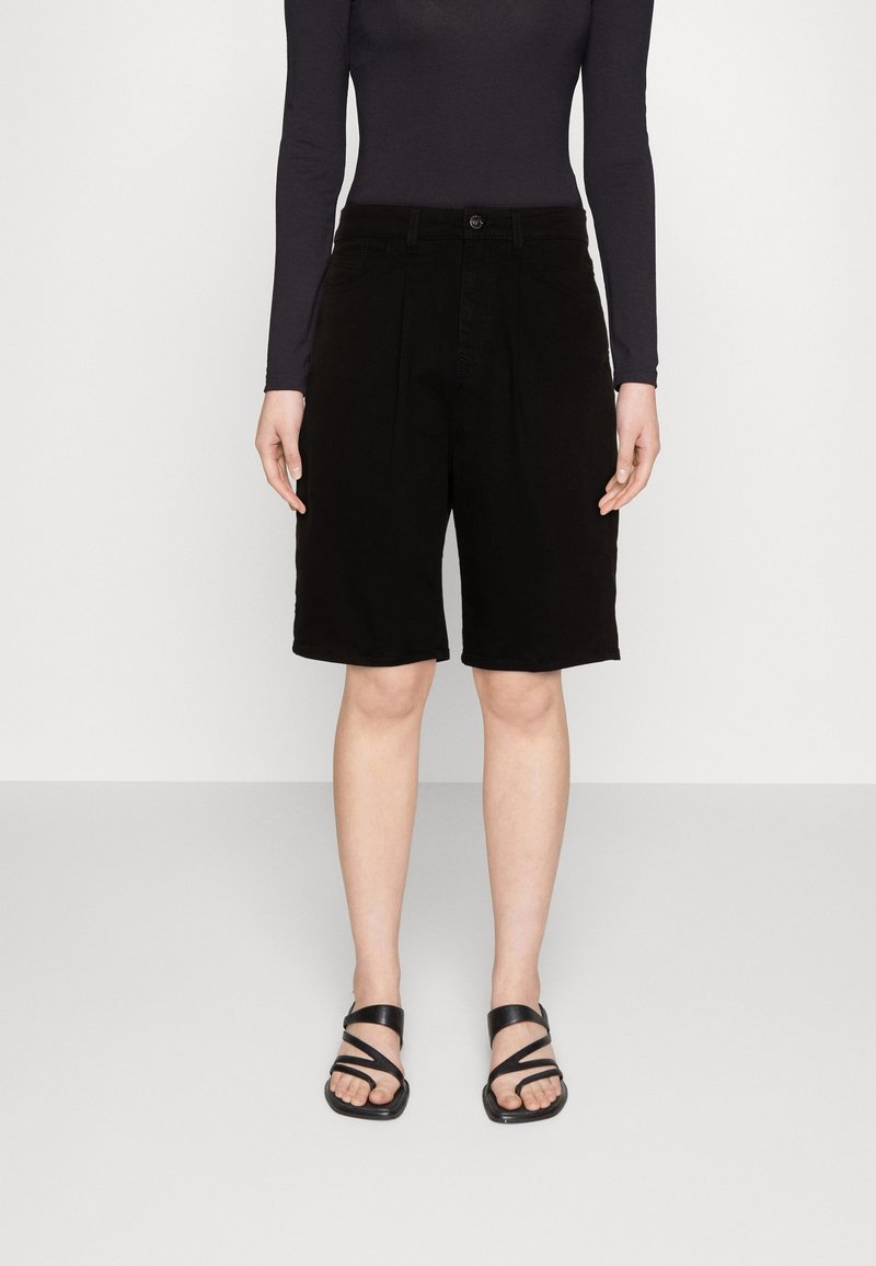 Henkilö, joka pukeutunut mustiin, vyötäröltä korkeavyötäröisiin polvipitkiin shortseihin, pitkähihainen tumma yläosa ja mustat remmityyliset tasapohjasandaalit, seisomassa yksivärisen taustan edessä.