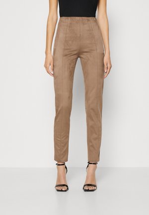 Vrouw draagt slanke, hoog uitgesneden bruine suède broek gecombineerd met zwarte open-teen enkellaarzen met bandje, staand tegen een effen achtergrond.