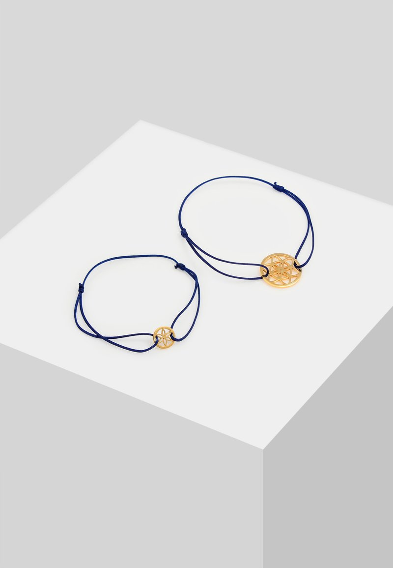 Pulseras de metal dorado con cordón azul, que presentan diseños circulares troquelados. Una pulsera es más grande y tiene una cuenta de acento; la otra es más pequeña.
