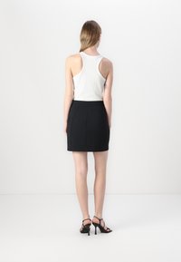 Sort tætsiddende nederdel med lynlås bagpå, parret med en hvid ribbet tanktop. Modellen står foran en ensfarvet baggrund og viser bagudsigten af outfittet.