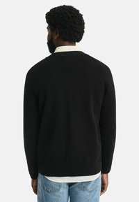 Schwarzer Strickpullover mit einem runden Ausschnitt und gerippten Bündchen, getragen über einem weißen Hemd; schlichtes Design und glatte Textur.