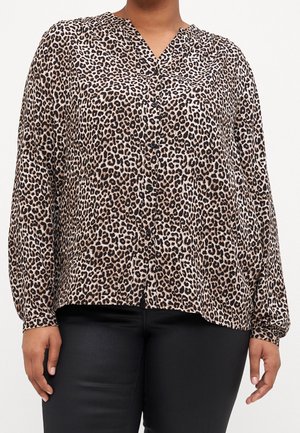 Personne portant un chemisier à manches longues à imprimé léopard boutonné, associé à un pantalon noir sur un fond uni.