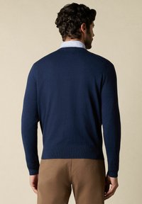 Marineblauer Pullover mit langen Ärmeln und geripptem Saum, aus weichem Stoff gefertigt. Über einem hellblauen Hemd mit Kragen getragen, von hinten gesehen.