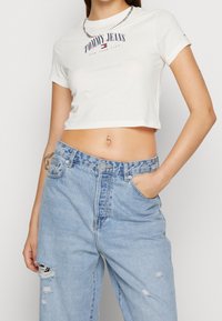 Bílý zkrácený tričko s logem "TOMMY JEANS", spárované se světle modrými roztrhanými vysokými džíny. Náhrdelník z řetízku jako doplněk.