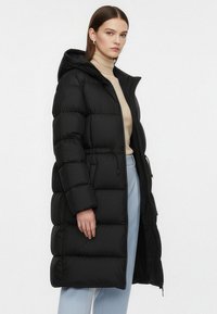 Sort dunjakke med hætte, med lodret quiltet design, justerbar snor i taljen og lynlåslukning foran.