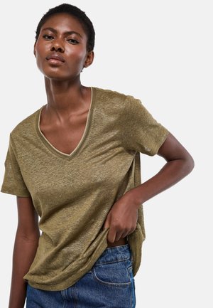 Femme aux cheveux courts portant un T-shirt olive à col en V et un jean bleu, posant avec une main dans la poche du jean.