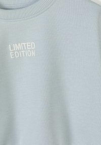 Vêtement bleu clair avec col côtelé présentant un texte brodé blanc "LIMITED EDITION" sur le côté gauche à l'avant.