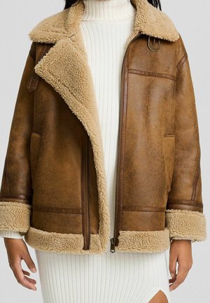 Veste en shearling marron avec doublure en fausse fourrure beige portée sur une robe blanche côtelée à col roulé avec fente latérale.