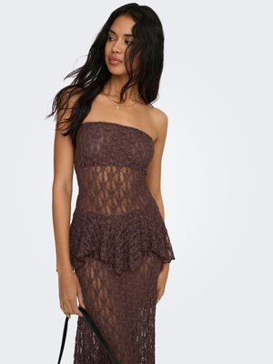 Femme portant une robe bustier en dentelle marron foncé avec un détail peplum, tenant une bretelle noire, regardant légèrement sur le côté sur un fond uni.