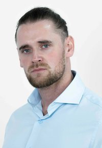 Close-up van een man met een lichtblauwe overhemdbloes, kort haar naar achteren gekamd en een baard. De achtergrond is vlak wit, waardoor zijn kenmerken worden benadrukt.