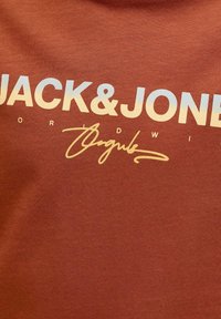 Ύφασμα σε χρώμα σκουριάς με λευκό κείμενο "JACK & JONES" και κίτρινη υπογραφή με καλλιγραφική γραφή κάτω από αυτή σε ένα κομμάτι ρούχου.