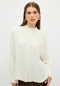 Crème-kleurige blouse met een hoge kraag, lange mouwen en een rechte snit. Glad textuur met een frontale knoopsluiting en subtiele glans.