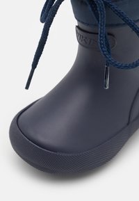 Bota de goma azul oscuro con suela texturizada, que cuenta con un puño de tela y cordones trenzados para un ajuste seguro. Acabado suave y brillante.
