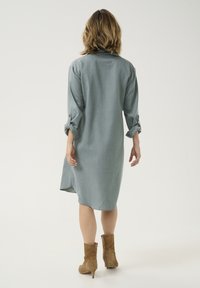 Robe grise clair, arrivée au genou, en tissu doux avec des manches longues retroussées aux coudes. Le dos présente un détail de pli subtil.
