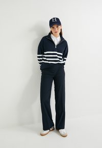 Marineblauwe hoodie met ritssluiting en witte horizontale strepen, gecombineerd met marineblauwe broek met wijde pijpen en witte sneakers. De pet heeft een logo.