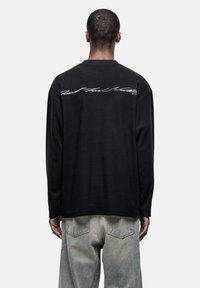Schwarzes Langarmshirt aus strukturiertem Stoff mit weißen geschwungenen Texten auf der oberen Rückseite. Kombiniert mit hellblauen Jeans.