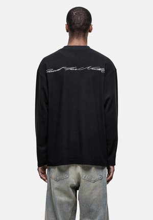 Zwarte long-sleeve shirt van een textuurstof, met witte cursieve tekst over de bovenrug. Gecombineerd met lichtblauwe denim jeans.