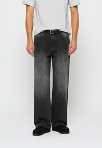 Tumši pelēki džinsi ar brīvu piegriezumu, pieci kabatas un smalka gradienta efekts. Valkāti ar pelēku T-kreklu un melnu apavu.