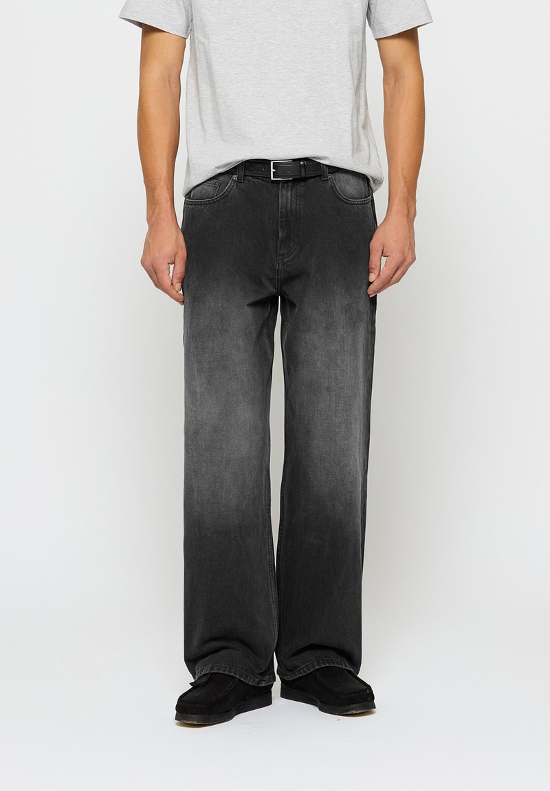 Tumši pelēki džinsi ar brīvu piegriezumu, pieci kabatas un smalka gradienta efekts. Valkāti ar pelēku T-kreklu un melnu apavu.