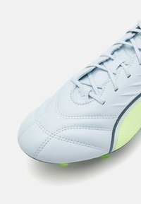 Scarpa da calcio azzurro chiaro con tomaia in pelle testurizzata, dettagli cuciti, tacchetti verdi e un accento giallo striato sulla suola.