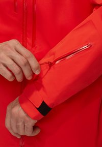 Rote wasserdichte Jacke mit einer Reißverschlusstasche am Ärmel, verstellbarem Bündchen mit Klettverschluss und glatter, glänzender Stoffoberfläche.