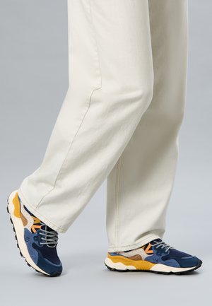 Persona che indossa pantaloni color crema e sneakers blu, beige e gialle su una superficie grigia.