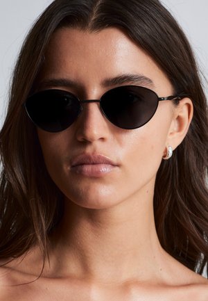 Vrouw met lang bruin haar, zwarte ovale zonnebril en kleine zilveren oorringen, neutrale uitdrukking, effen lichte achtergrond.