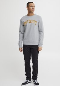 Sudadera gris con "UNIVERSIDAD" en texto amarillo en negrita. Combinada con jeans negros y zapatillas negras. Diseño minimalista, cuello redondo.