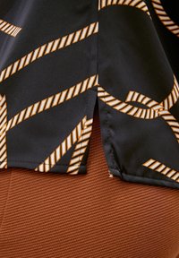 Chemise en tissu noir avec un motif de rayures diagonales orange et blanches, dotée d'une ourlet arrondi. Un bas texturé marron est visible.