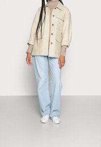 Veste-chemise beige clair avec des boutons, deux poches avant et manches retroussées, associée à un jean bleu clair à jambes larges et des baskets blanches.