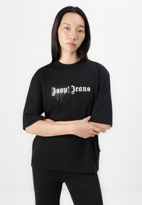 Melna kokvilnas T-kreklā ar īsām piedurknēm, uz kura ir balts teksts "Joop Jeans" stilizētā fontā, papildināts ar izkaisītiem baltiem elementiem.
