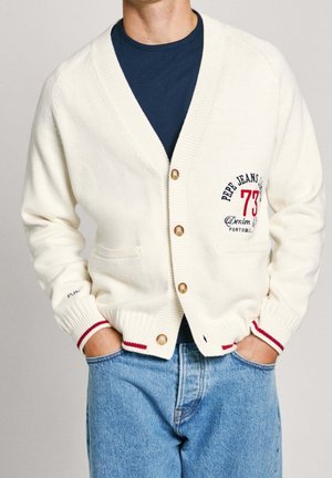 Homme portant un cardigan blanc boutonné avec un logo rouge et bleu et un jean bleu clair, les mains dans les poches du jean.