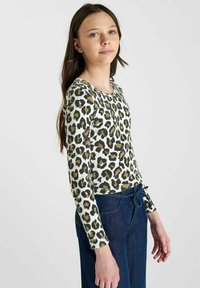 Langsleeve t-shirt van ribfluwelen stof met een witte basis en een groene en zwarte luipaardprint. Gecombineerd met blauwe denim broek met een trekkoord.