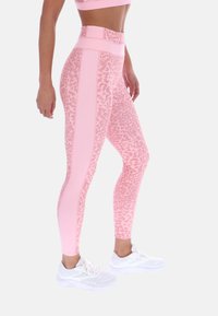 Γυναίκα που φοράει ροζ κολάν λεοπάρ με ψηλή μέση και μονόχρωμες πλαϊνές ρίγες, καθώς και λευκά αθλητικά παπούτσια, στέκεται πλευρικά.